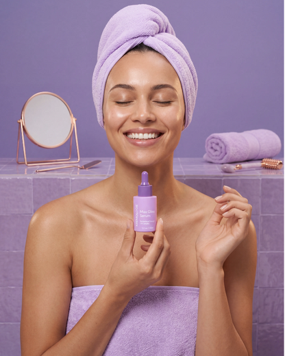 Woman applying KREMOLOGIE MAX GLOW SERUM skincare product with a purple towel and mirror in the background  
